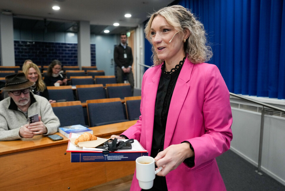 Digitaliserings- og forvaltningsminister Karianne Tung har sendt et forslag til norsk AI-lov på høring. Bildet er av en formelt kledd Karianne Tung som bærer en kopp i en hånd, og en perm og dokumenter i den andre. Hun har en bøylemikrofon på, og er inne i en sal der folk sitter på rekke.
