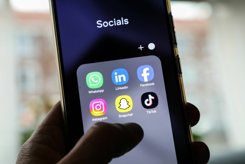 Annonser som er mikset inn blant vanlige meldinger kan være ulovlig, ifølge Forbrukertilsynet. De mener disse annonsene kan rammes av spam-lovverket. Bildet er av en hånd som holder en smarttelefon. Tommelen 'svever' over skjermen, og skjermbildet viser en mappe med logoene til ulike sosiale medier.