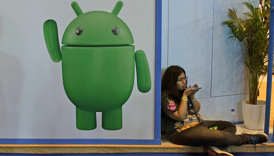 Android-utviklere skal få bedre AI-hjelp i Android Studio. En kvinne sitter på gulvet på en Comic Con-konferanse, med en stor Android-logo på veggen ved siden av.