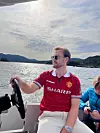 Bildet er av en ung mann bak rattet på en båt. Han har lys bukse, solbriller og rød Manchester United-fotballdrakt. I bakgrunnen ser vi sjø.
