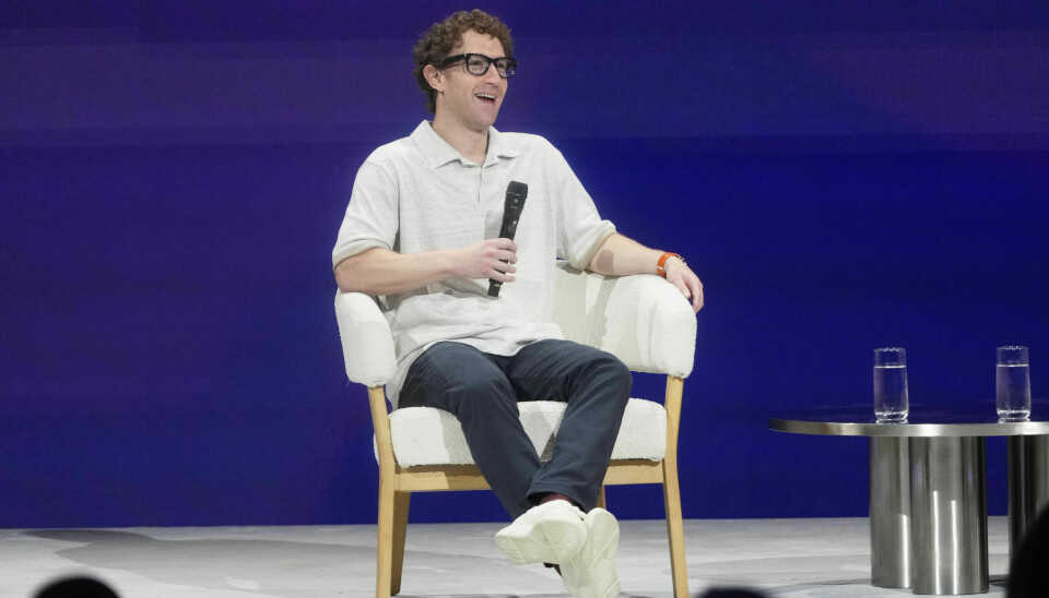 Du bør være forsiktig med å dele personlig eller sensitiv informasjon på Messenger-chat, advarer Meta. Innhold i chatter kan nemlig brukes til å trene Meta og Mark Zuckerbergs AI. Bildet er av en mann i hvit skjorte og hvite sko, med mørk bukse og mørke briller med tjukk innfatning. Han sitter i en hvit stol, på en hvit scene foran en blå vegg og holder en mikrofon. Mannen er Mark Zuckerberg.