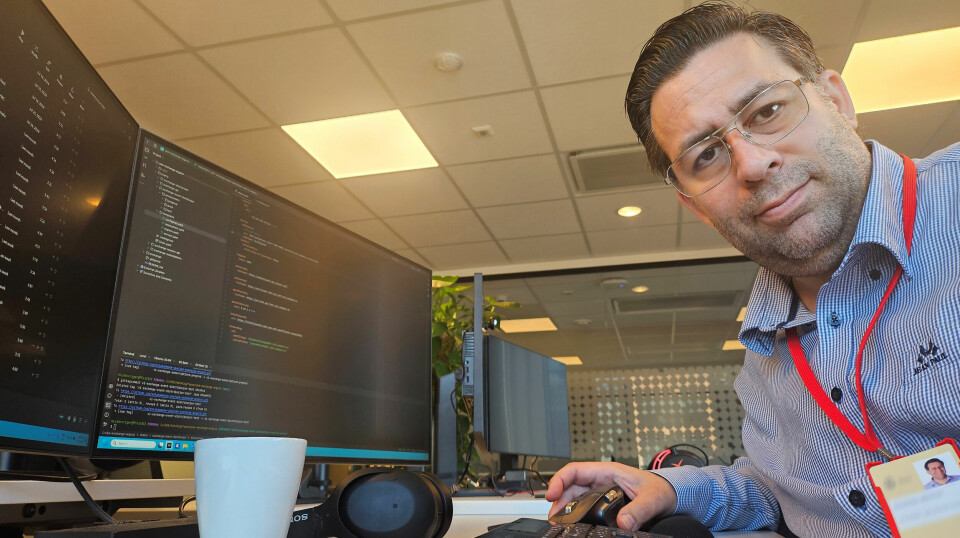 Cristhian Gertner mener IT-bransjen må tørre å ansette flere unge. – Dette er ikke veldedighet. Det er den mest kritiske og lønnsomme investeringen vi kan gjøre i vår felles fremtid, mener han. Bildet er av en mann som ser i kamera. Han har på skjorte, og rundt halsen har han et rødt bånd med nøkkelkort. Han sitter ved en pult der det er to skjermer med kode.